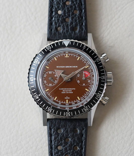 A Nivada Grenchen Chronomaster Broad Arrow watch with a black racing strap and stainless steel case, featuring a matte black dial with cream colored luminous indexes and shiny silver hands.